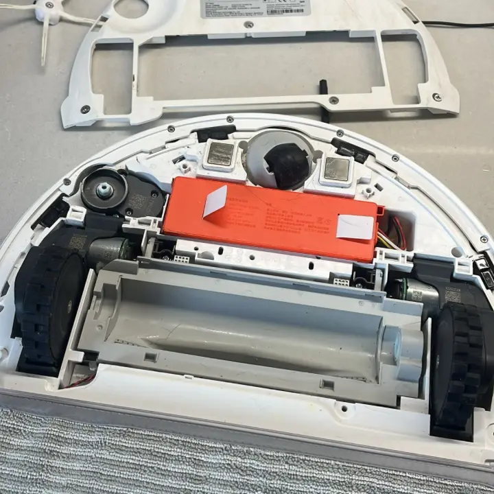 Atvērts Xiaomi robottīrītājs, redzama sarkanā baterija, birste noņemta.